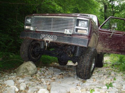 1980 Ford Bronco - A maroon Ford pickup truck with large off-road tires is navigating a rocky terrain with its driver's side door open. The truck's front bump…