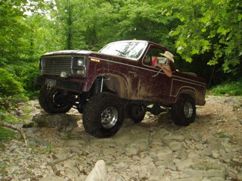 1980 Ford Bronco - A lifted, maroon Ford Bronco is navigating a rocky, wooded trail. The driver, wearing a cowboy hat, is visible through the driver's side wi…