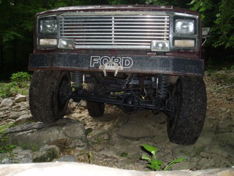 1980 Ford Bronco - The front view of a lifted, dark red 1980 Ford Bronco is shown navigating a rocky, wooded terrain. The vehicle's front grille, headlights,…