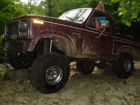 1980 Ford Bronco - A lifted, maroon 1980 Ford Bronco is navigating a rocky, off-road trail. Mud is splattered across the vehicle's exterior, and a person is v…