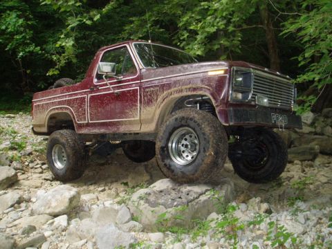 1980 Ford Bronco - A lifted, maroon Ford Bronco is navigating a rocky, wooded trail. The vehicle is covered in mud, indicating off-road use.