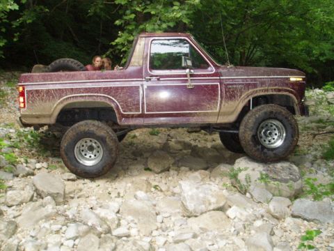 1980 Ford Bronco - A maroon 1980s Ford Bronco is navigating a rocky, off-road trail. The vehicle is lifted and has large tires, with two people visible in the…