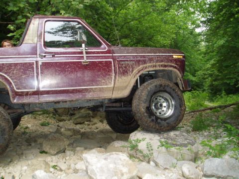 1980 Ford Bronco - A maroon Ford Bronco is shown from the side, navigating a rocky trail with mud splattered on its body. The vehicle is off-roading through a…