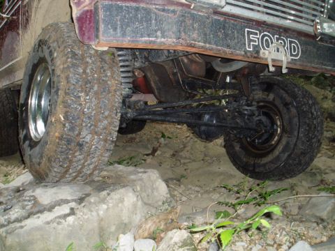 1980 Ford Bronco - The undercarriage and rear wheel of a dark red Ford Bronco are visible, with the front of the vehicle showing the word "FORD" in white lett…