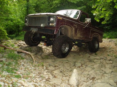 1980 Ford Bronco - A lifted, maroon Ford Bronco is navigating a rocky, wooded trail. The front of the vehicle displays the "FORD" logo on the bumper.