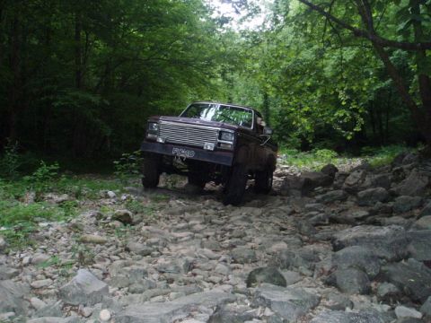 1980 Ford Bronco - A maroon Ford pickup truck navigates a rocky, wooded trail. The truck's front grille and headlights are clearly visible as it drives over u…