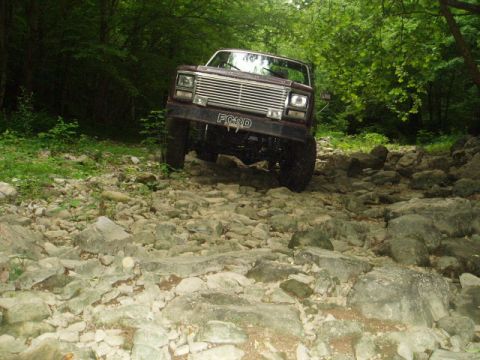 1980 Ford Bronco - A maroon Ford Bronco is navigating a rocky, wooded trail. The front grille of the vehicle is clearly visible with the word "FORD" embossed…