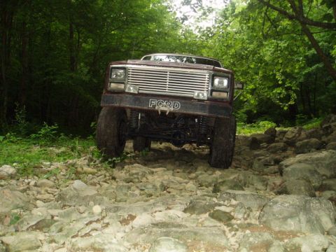 1980 Ford Bronco - A lifted, dark red Ford pickup truck with a chrome grille navigates a rocky, wooded trail. The front of the vehicle is angled towards the c…