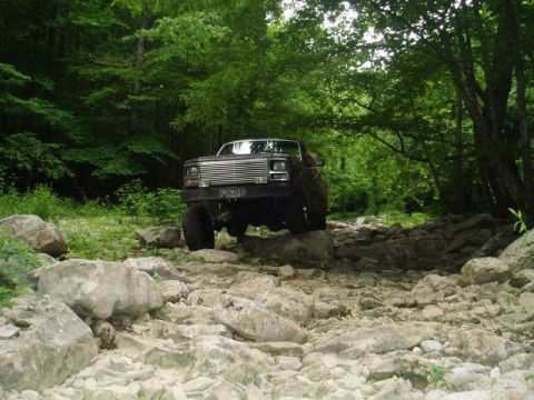 1980 Ford Bronco - A dark red 1980s era pickup truck with a lifted suspension navigates a rocky, wooded trail. The truck has a chrome grille and a license pla…