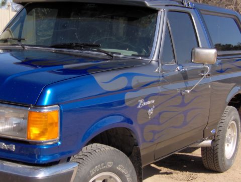 1987 Ford Bronco - The front quarter panel and hood of a blue and gray Ford Bronco with flame graphics are visible. The vehicle has a chrome bumper and an ora…