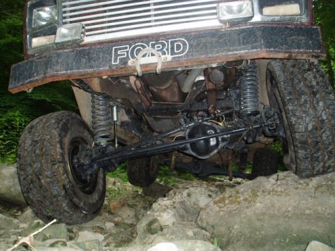 1980 Ford Bronco - The underside of a lifted Ford Bronco is shown navigating a rocky trail. The front suspension, differential, and tires are visible, with th…