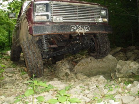1980 Ford Bronco - A maroon Ford Bronco is navigating a rocky, off-road trail with its front tires angled upwards over large rocks. The front grille and bumpe…