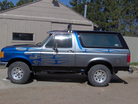 1987 Ford Bronco - A side view of a customized gray and blue Ford Bronco with flame graphics parked in front of a building. The vehicle has a black roof and t…