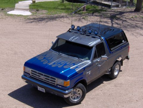 1987 Ford Bronco - A custom blue and silver Ford Bronco is parked on a gravel surface, featuring flame decals on the hood and sides, a roof-mounted light bar,…