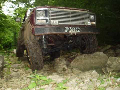 1980 Ford Bronco - A lifted, maroon Ford pickup truck is navigating a rocky, wooded trail. The front of the truck is angled towards the camera, with its large…