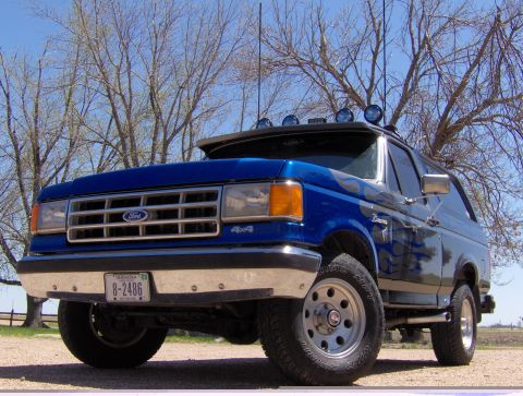 1987 Ford Bronco - A blue 1987 Ford Bronco with flame graphics is parked outdoors under a clear blue sky. The front of the SUV is visible, showing a chrome gr…