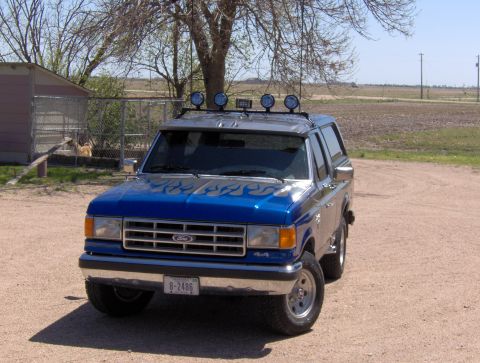 1987 Ford Bronco - A blue 1987 Ford Bronco with flame decals on the hood is parked on a dirt road in front of a small building and a fenced area. The vehicle…