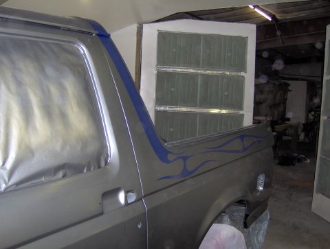 1987 Ford Bronco - The side of a dark gray Ford Bronco is shown, featuring blue flame decals on the rear fender and masking tape around the windows. The vehic…