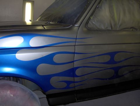 1987 Ford Bronco - A close-up side view of a vehicle's fender and door featuring a custom blue and silver flame graphic. The vehicle appears to be in a worksh…