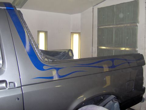 1987 Ford Bronco - The side of a silver Ford Bronco with blue flame graphics is shown in a spray booth. The vehicle has a custom blue stripe running up the re…