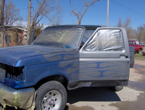 1987 Ford Bronco - A blue and silver Ford truck is being prepped for paint, with flames painted on the side. The windows and front end are covered in plastic…
