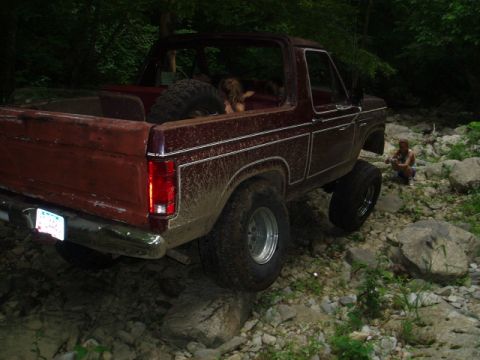 1980 Ford Bronco - A maroon Ford Bronco is navigating a rocky, wooded terrain, with its tires positioned on large stones. A person is visible crouching in the…