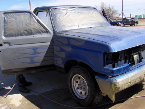 1987 Ford Bronco - A blue Ford pickup truck with flame decals is being prepped for painting, with the driver's side door open and covered in plastic.