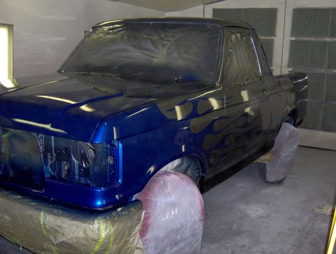 1987 Ford Bronco - A blue pickup truck with flames painted on the side is in a paint booth. The front of the vehicle is visible, and the wheels are covered.
