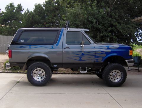 1987 Ford Bronco - A lifted, two-tone gray and blue 1987 Ford Bronco with flame graphics is parked on a concrete driveway in front of trees.