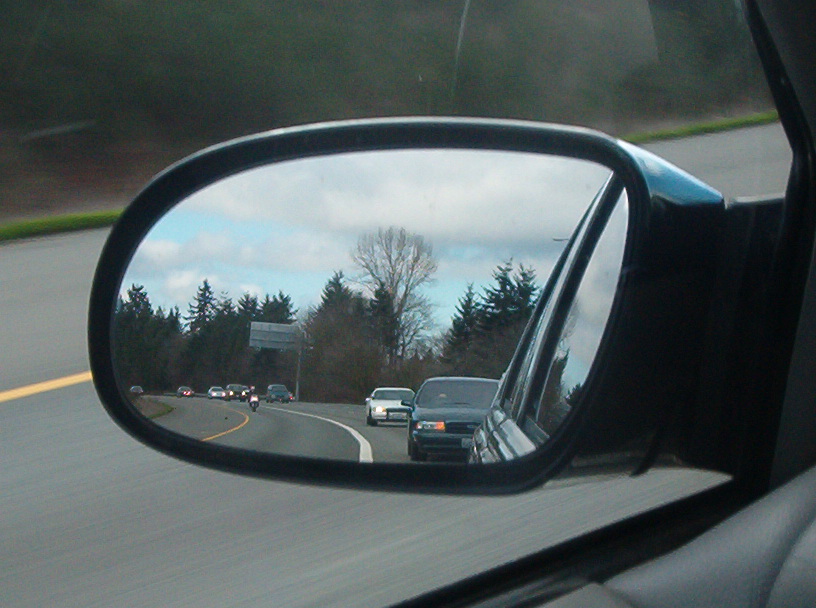 Driver's side mirror reflecting a line of following traffic on a tree-lined highway