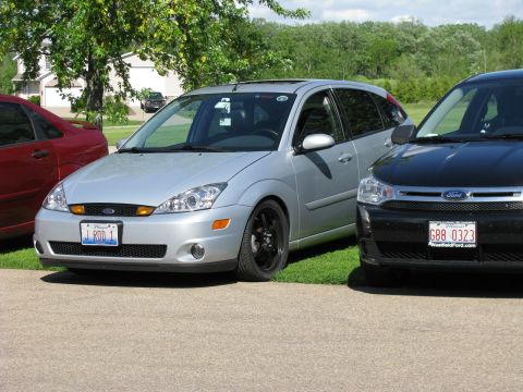 2003 Ford Focus - A silver 2003 Ford Focus hatchback is parked next to a black Ford Focus. The silver Focus has a custom license plate that reads "J ROD 1".