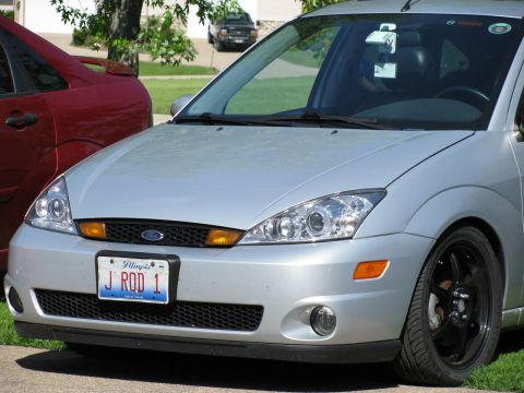 2003 Ford Focus - Front view of a silver Ford Focus with a custom license plate reading "J ROD 1" and black aftermarket wheels. A red car is partially visibl…