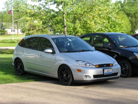2003 Ford Focus - A silver 2003 Ford Focus hatchback is parked on a paved surface in front of a grassy area and trees. The car has black aftermarket wheels a…