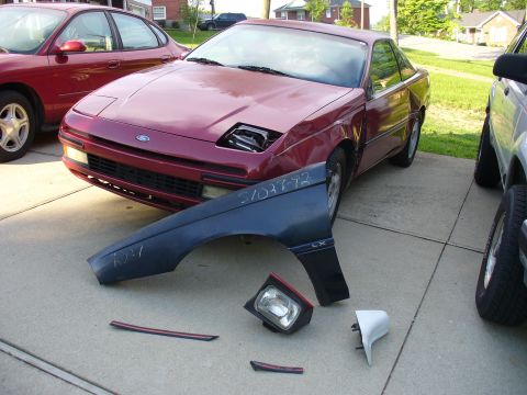 1991 Ford Probe - A maroon Ford Probe sits in a residential driveway mid-teardown with its front fender and passenger pop-up headlamp removed, exposing the e…