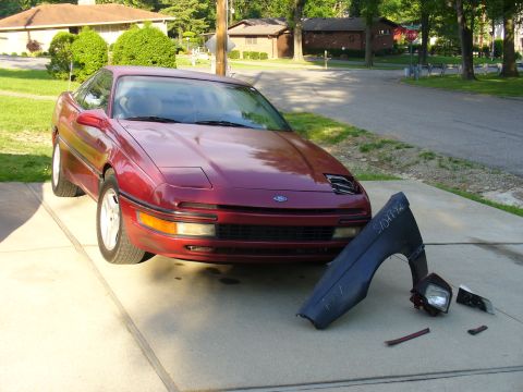 1991 Ford Probe - Maroon first-generation Ford Probe on a residential driveway with its right front fender removed, exposing the open headlight-pod cavity; t…