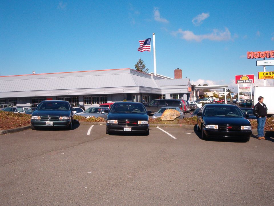 Two black Impala SS sedans and a green B-body Chevy parked in front of a Hooters restaurant