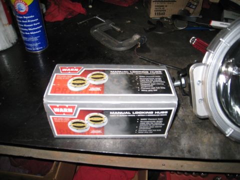 1996 Ford Bronco - A box of two Warn manual locking hubs sits on a dark workbench next to a large, round auxiliary light. The box has red and silver branding…