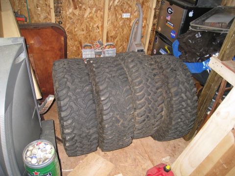 1996 Ford Bronco - Four large off-road tires are leaning against a wall in a cluttered garage. A silver CRT television is visible to the left, and a red gas c…