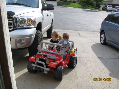 1994 Ford Bronco - Two children sit in a red toy Jeep, with a large pickup truck to their left and a blue car to their right.