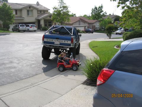 1994 Ford Bronco - A lifted Chevrolet pickup truck with a roll cage is parked behind a red toy jeep carrying two children.