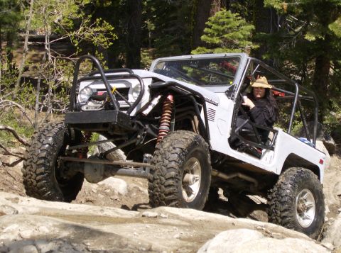 1986 Ford Bronco - A modified white off-road vehicle with large tires is navigating a rocky trail. A person wearing a hat is visible in the driver's seat.