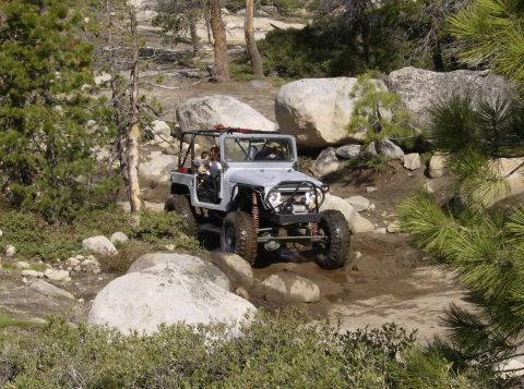 1986 Ford Bronco - A lifted, light gray Jeep-style off-road vehicle navigates a muddy, rocky trail with large boulders and pine trees surrounding it.