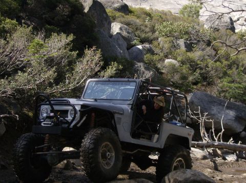 1986 Ford Bronco - A modified gray off-road vehicle with a roll cage is parked on a rocky, wooded trail. A person wearing a hat is visible in the driver's sea…