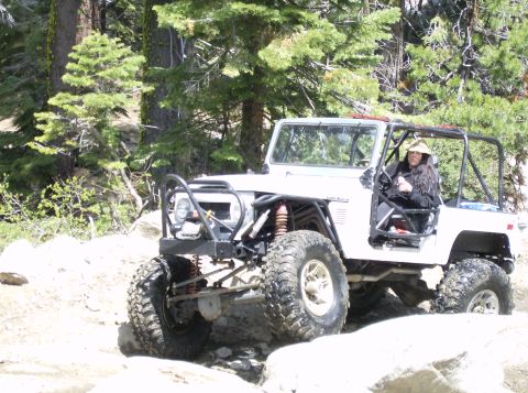 1986 Ford Bronco - A white off-road SUV with large tires is navigating a rocky trail surrounded by trees. The driver, wearing a hat, is visible behind the whe…