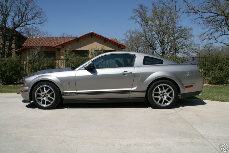 Silver Shelby GT500 Mustang coupe, side view, with dark rocker stripes on a driveway.