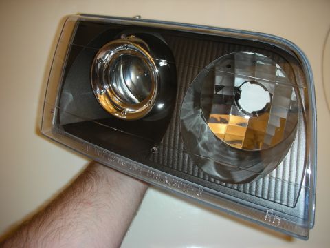 1998 Ford Crown Victoria - hand holds the front of a black-housing projector headlight with a chrome projector and clear lens.