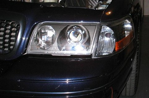 1998 Ford Crown Victoria - Close-up of a dark car's front corner with an illuminated aftermarket projector headlight and amber turn signal.