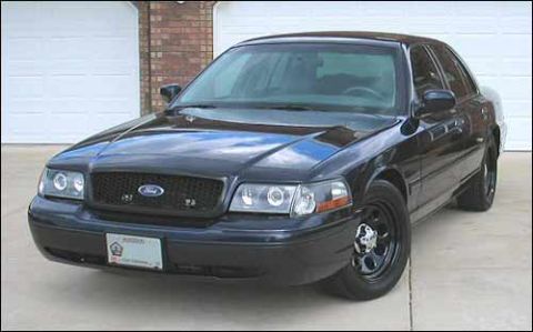 1998 Ford Crown Victoria - Front three-quarter view of a dark blue Ford full-size sedan with black wheels parked by a brick garage.