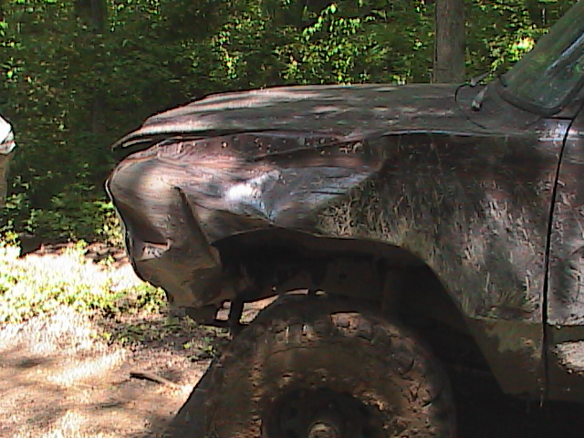 Mud-covered pickup truck with damaged front fender and hood, parked in a wooded area