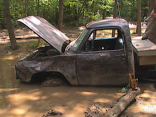 Mud-covered truck with hood open stuck in muddy water, trees in background